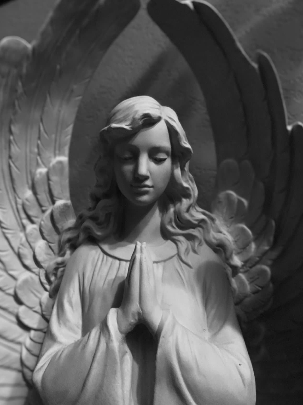 Un ángel esculpido con las manos juntas y la mirada baja, con las alas elevándose detrás.